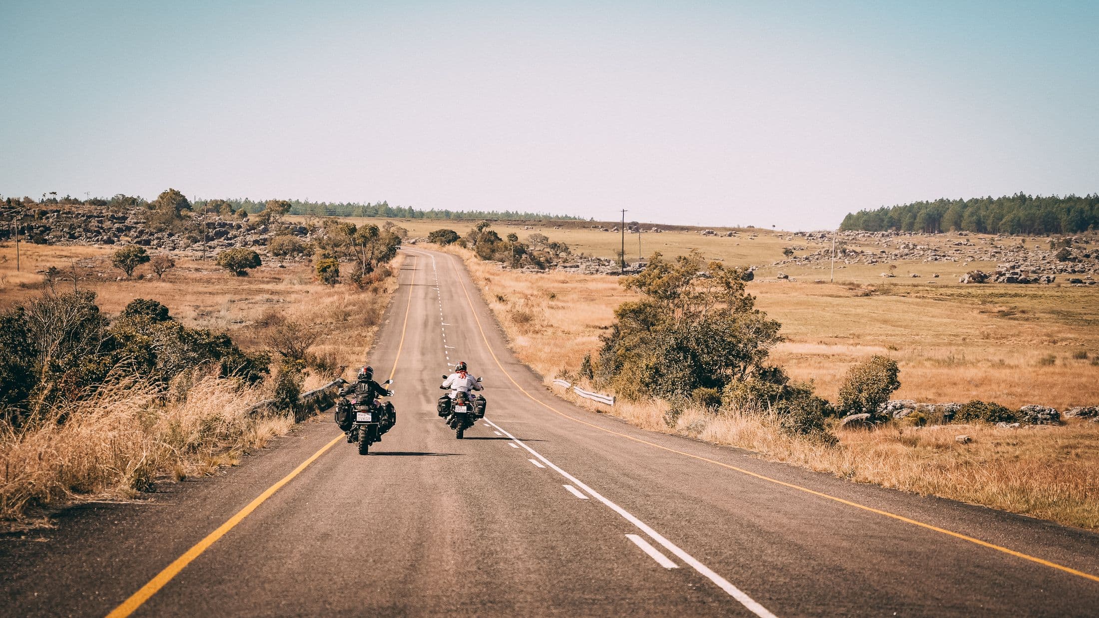 Two riders on an open South African road at golden hour, following the yellow edge line